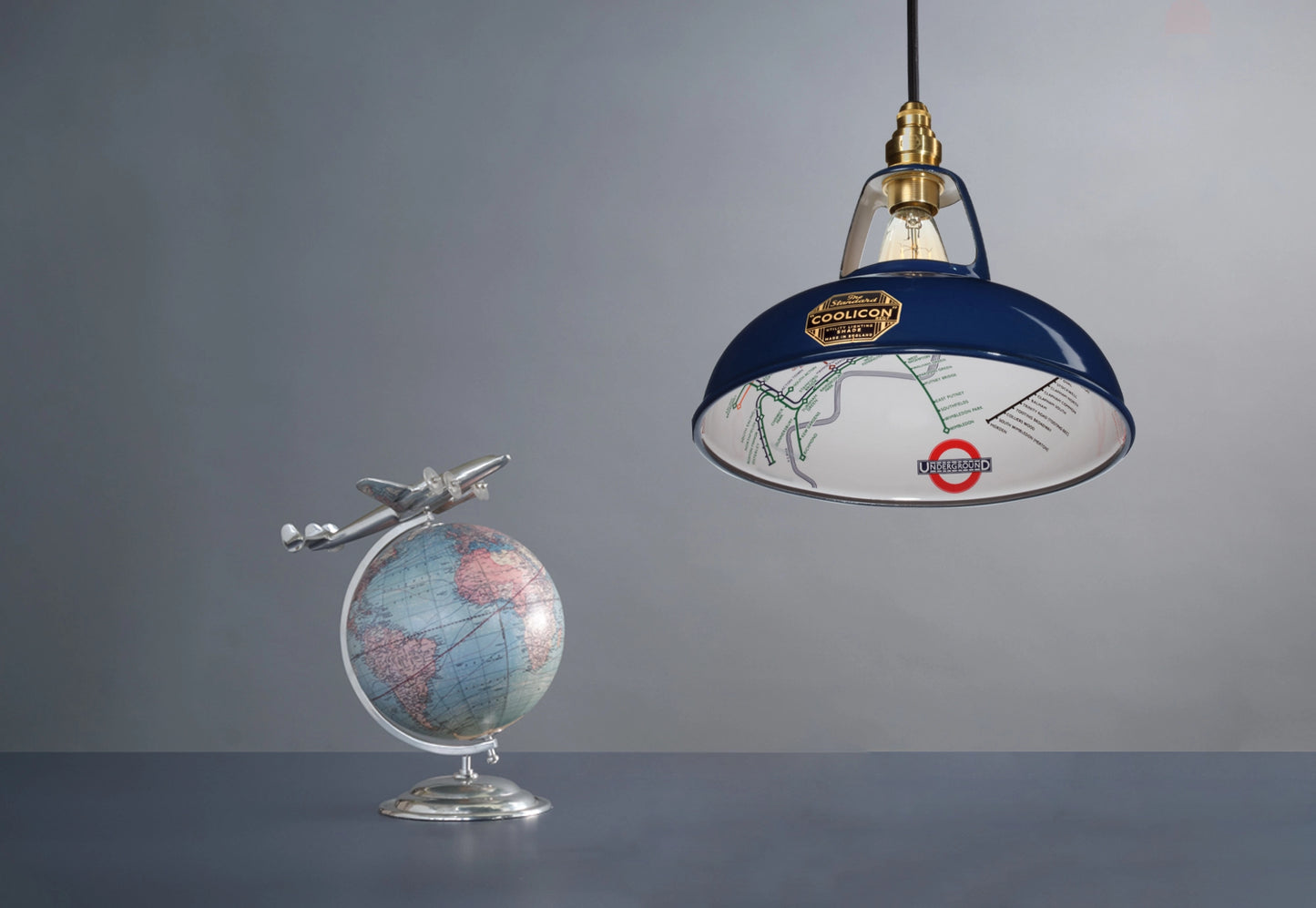 An Original Piccadilly Line Blue shade over a table. On its left is a globe with a metal plane attached to the top.