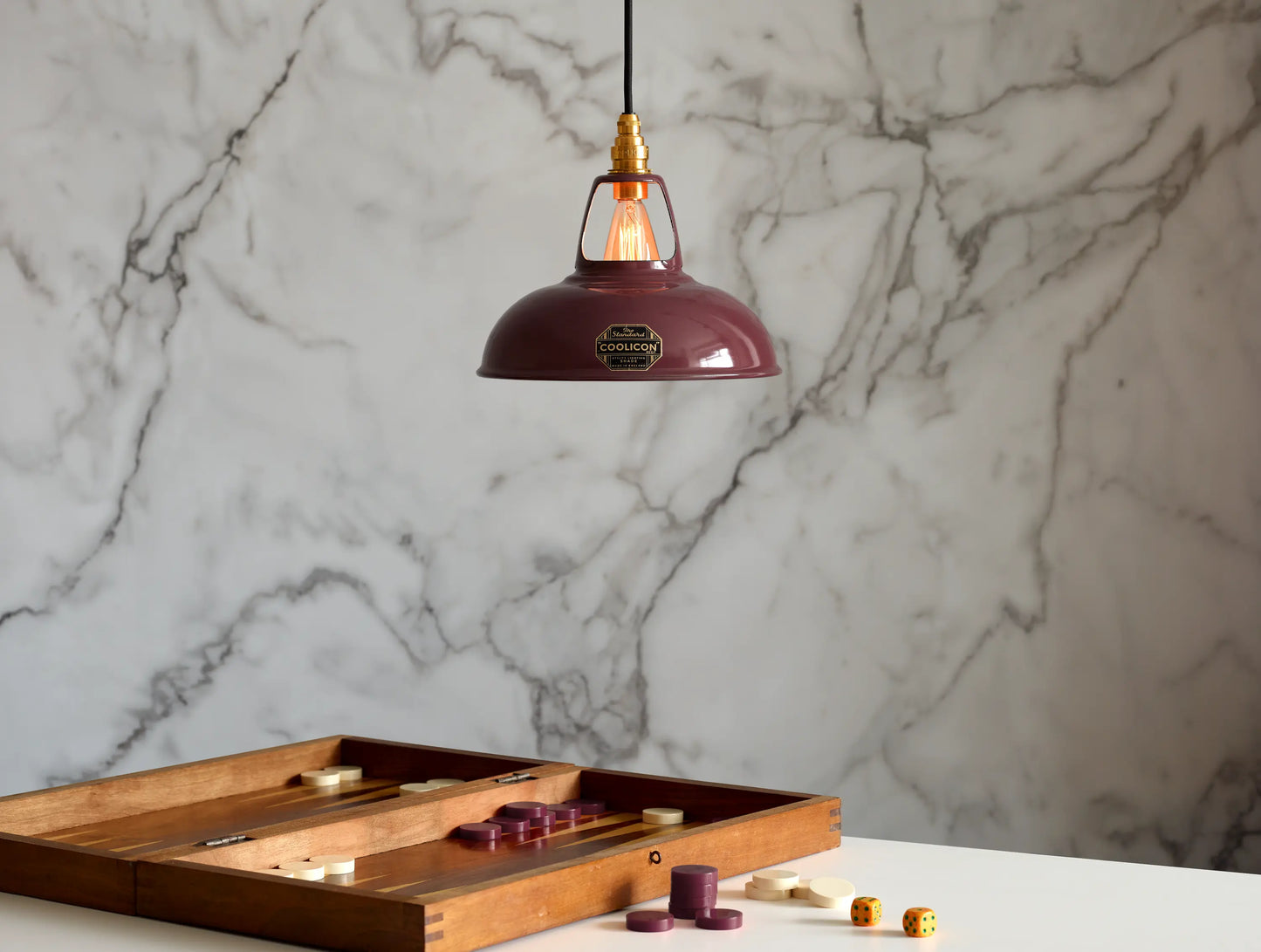 A Coolicon Metropolitan burgundy lampshade with a Signature Brass pendant set hanging above a table with an opened game of Backgammon. The background is a marble wallpaper.