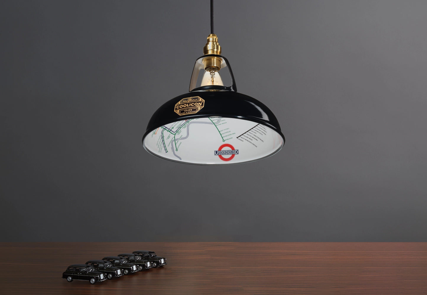 An Original Northern Line Black shade hanging over a wooden table with a line of toy cars of black taxis.