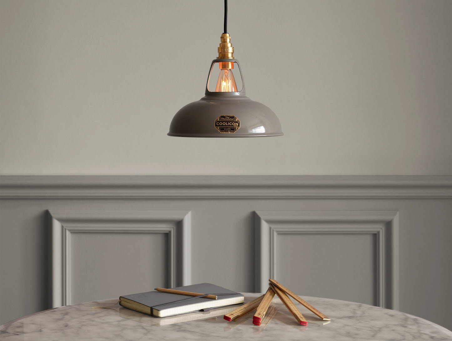 A Coolicon Original Grey lampshade hanging over a marble table. Below there are a grey notebook, an orange pencil and a wooden folding ruler 