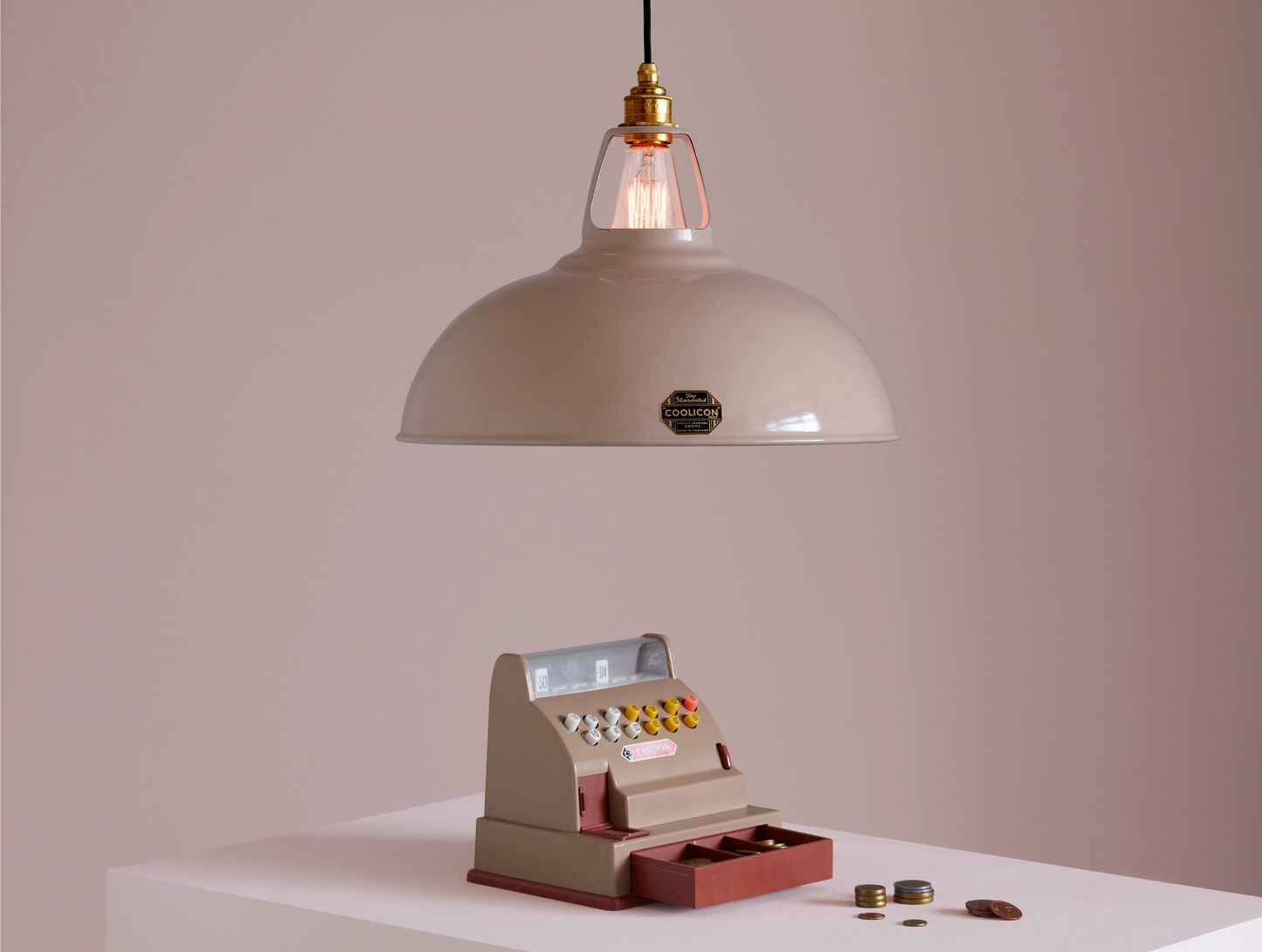 A Large Coolicon Latte Brown lampshade with a Porcelain pendant set hanging above a plinth with a vintage-looking light brown toy cash register and coins