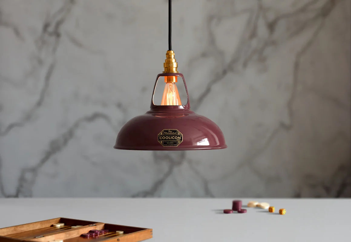 A Coolicon Metropolitan burgundy lampshade with a Signature Brass pendant set hanging above a table with an opened game of Backgammon. The background is a marble wallpaper.