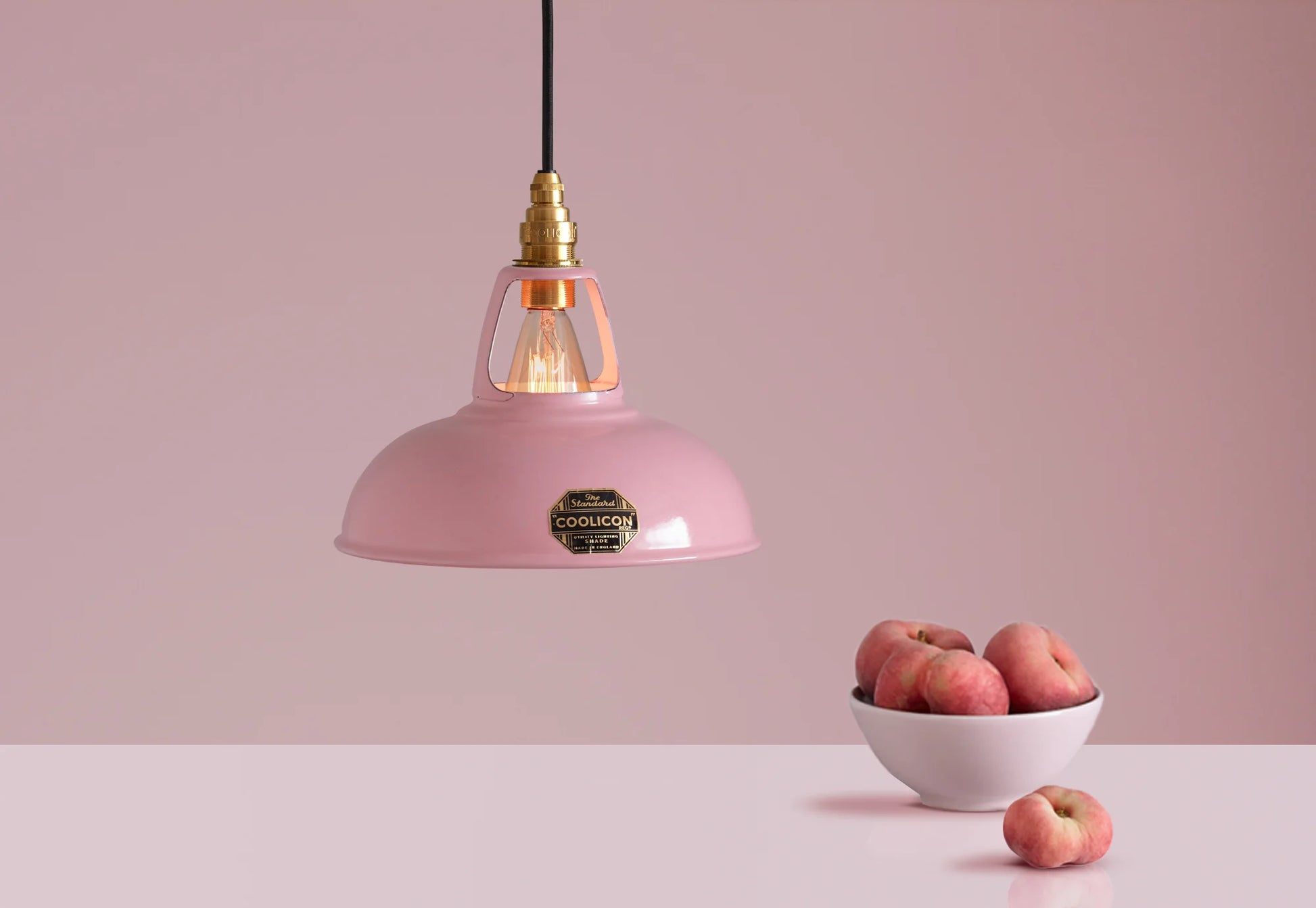 A Coolicon Powder Pink lampshade with a Brass pendant set hanging above a plinth with a bowl of flat peaches.