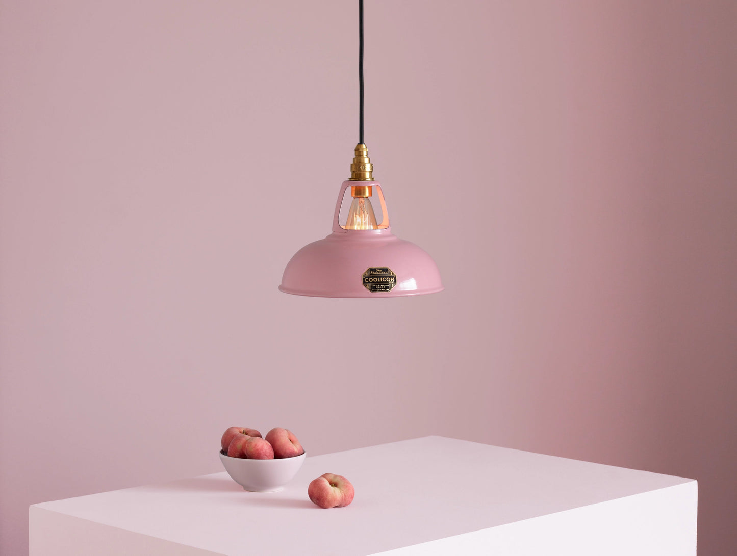 A Coolicon Powder Pink lampshade with a Brass pendant set hanging above a plinth with a bowl of flat peaches.