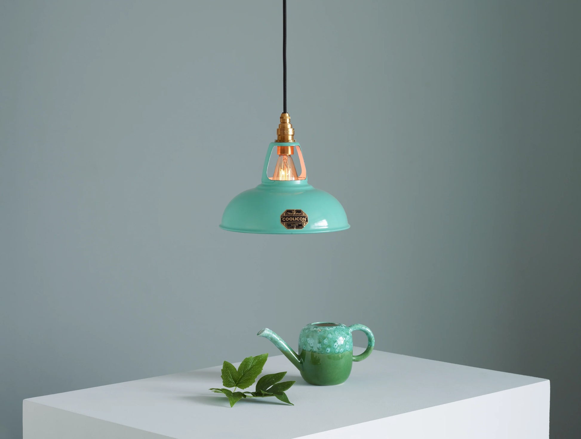 A Coolicon Fresh Teal lampshade hanging over a plinth. Below the shade is a large teal teapot and a branch with green leaves