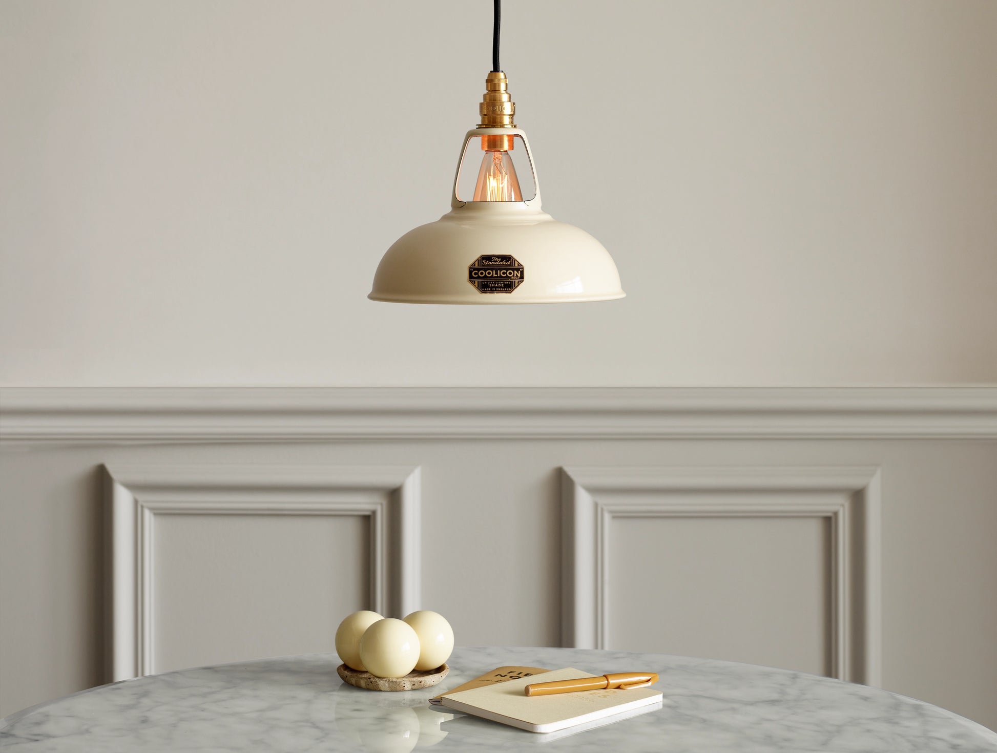 A Coolicon Classic Cream lampshade hanging over a marble table. Three cream snooker balls, two notebooks and an orange pen are placed on the table