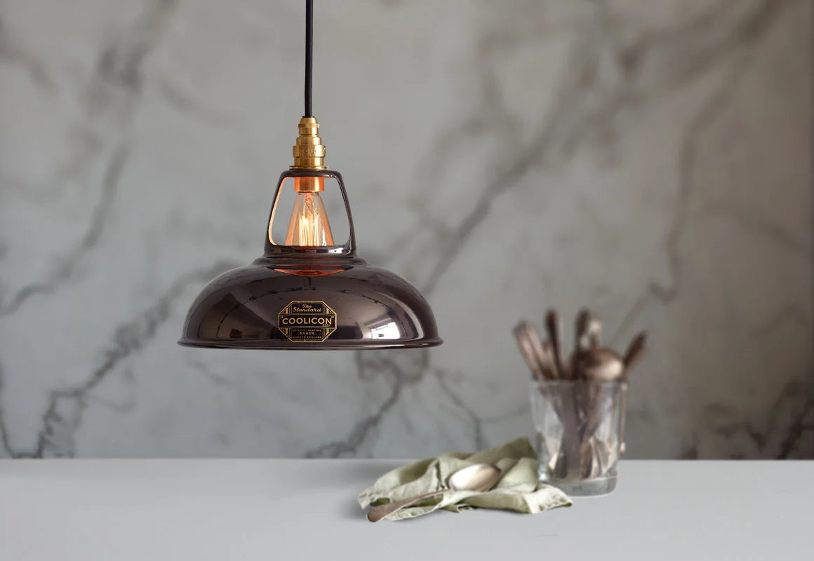 A Coolicon Pewter grey lampshade with a Signature Brass pendant set hanging above a table with a glass container with a set of old cutlery. The background is a marble wallpaper.