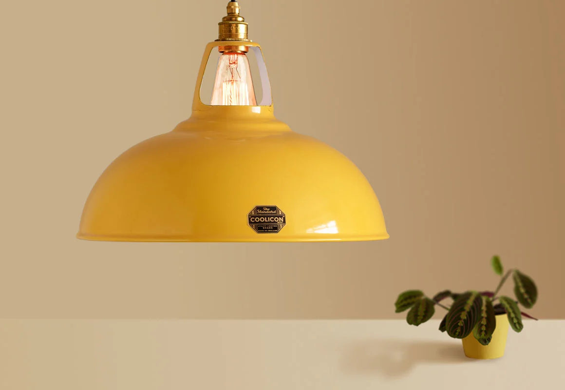 A Large Coolicon Deep Yellow lampshade hanging over a plinth. Below the shade is a pinstripe calathea plant in a yellow plant pot 