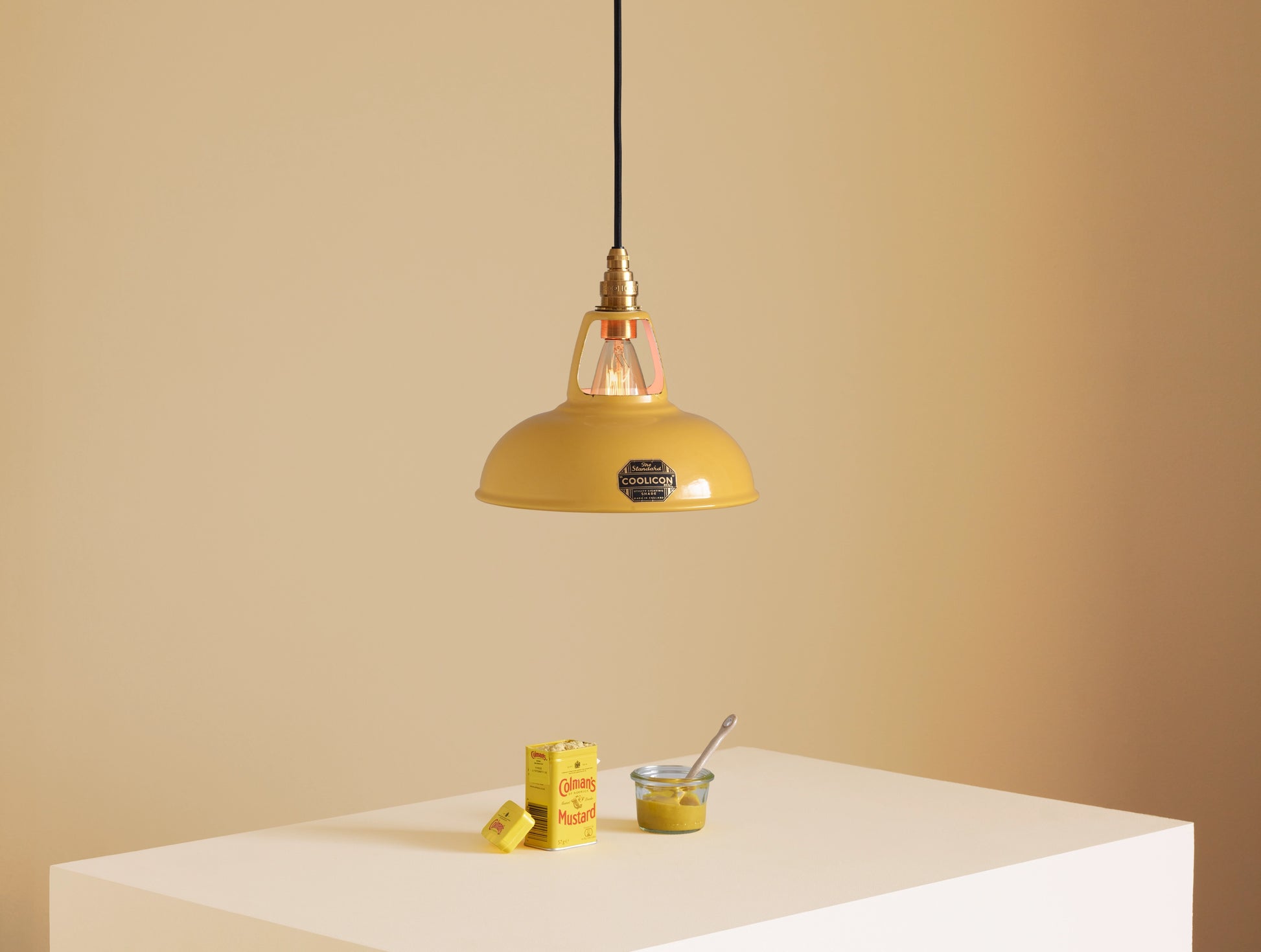 A Coolicon Deep Yellow lampshade hanging over a plinth. Below the shade is a tin of Colman’s powder mustard, and a glass jar with mustard in it