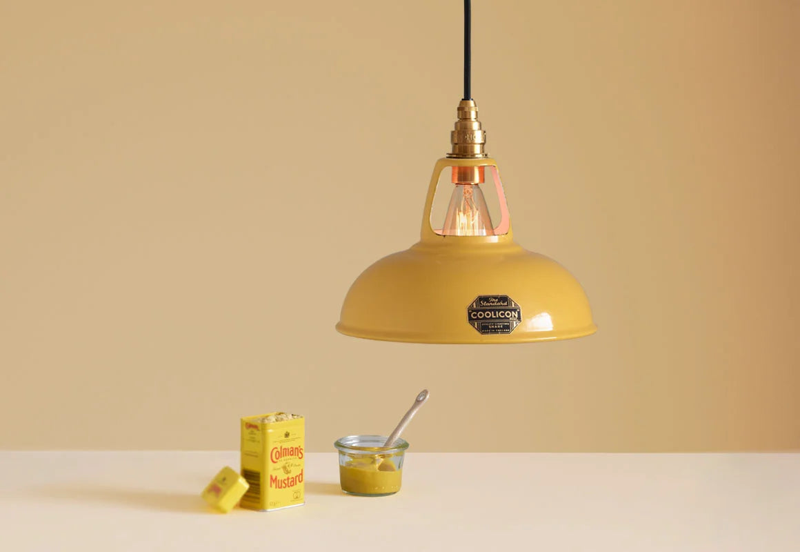 A Coolicon Deep Yellow lampshade hanging over a plinth. Below the shade is a tin of Colman’s powder mustard, and a glass jar with mustard in it