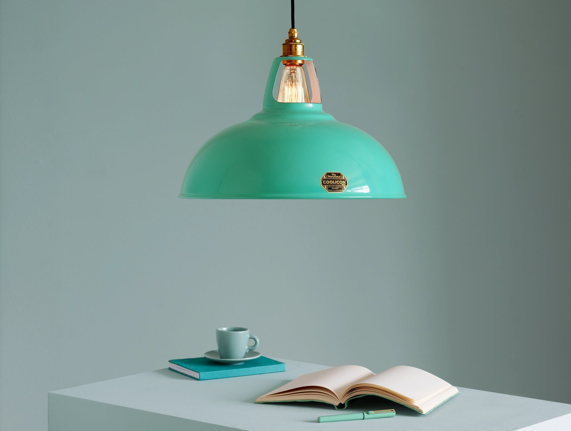 A Large Coolicon Fresh Teal lampshade hanging over a plinth. Below the shade is an open notebook, a teal pen, and a teal espresso cup and saucer on top of a closed teal notebook
