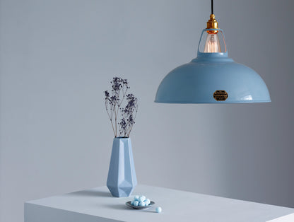 A Large Coolicon Sky Blue lampshade with a Brass pendant set hanging above a plinth with a bouquet of dried broom blue flowers inside a blue vase. On its side there is a small plate with blue bonbons