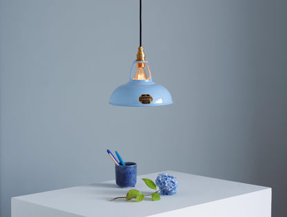 A Coolicon Sky Blue lampshade with a Brass pendant set hanging above a plinth with a blue hydrangea flower and a dark blue pencil cup containing two blue pens