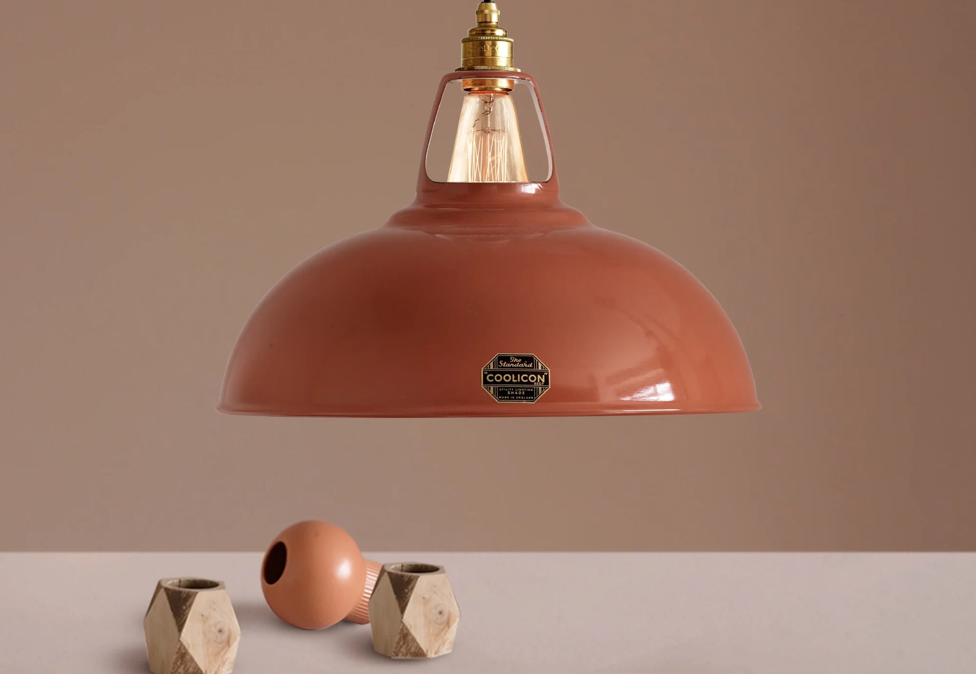 A Large Coolicon Terracotta lampshade hanging over a plinth. Below the shade is a small orange flower vase and a small wooden vase