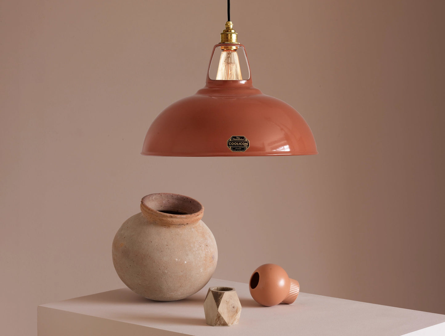 A Large Coolicon Terracotta lampshade hanging over a plinth. Below the shade is a large terracotta amphora, a small orange flower vase and a small wooden vase