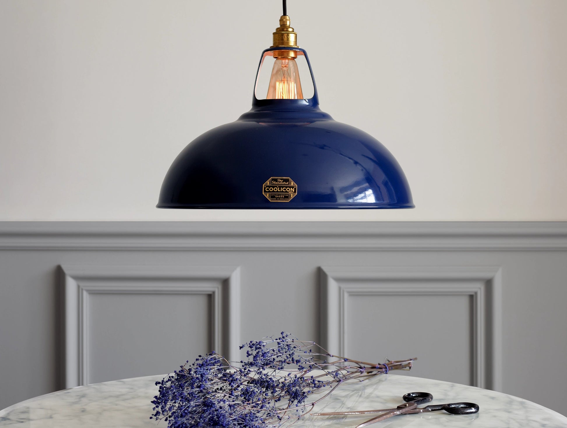 A large Royal Blue Coolicon shade hanging over a marble table. There is a branch of dried broom blue flowers underneath, along with a rusty pair of scissors