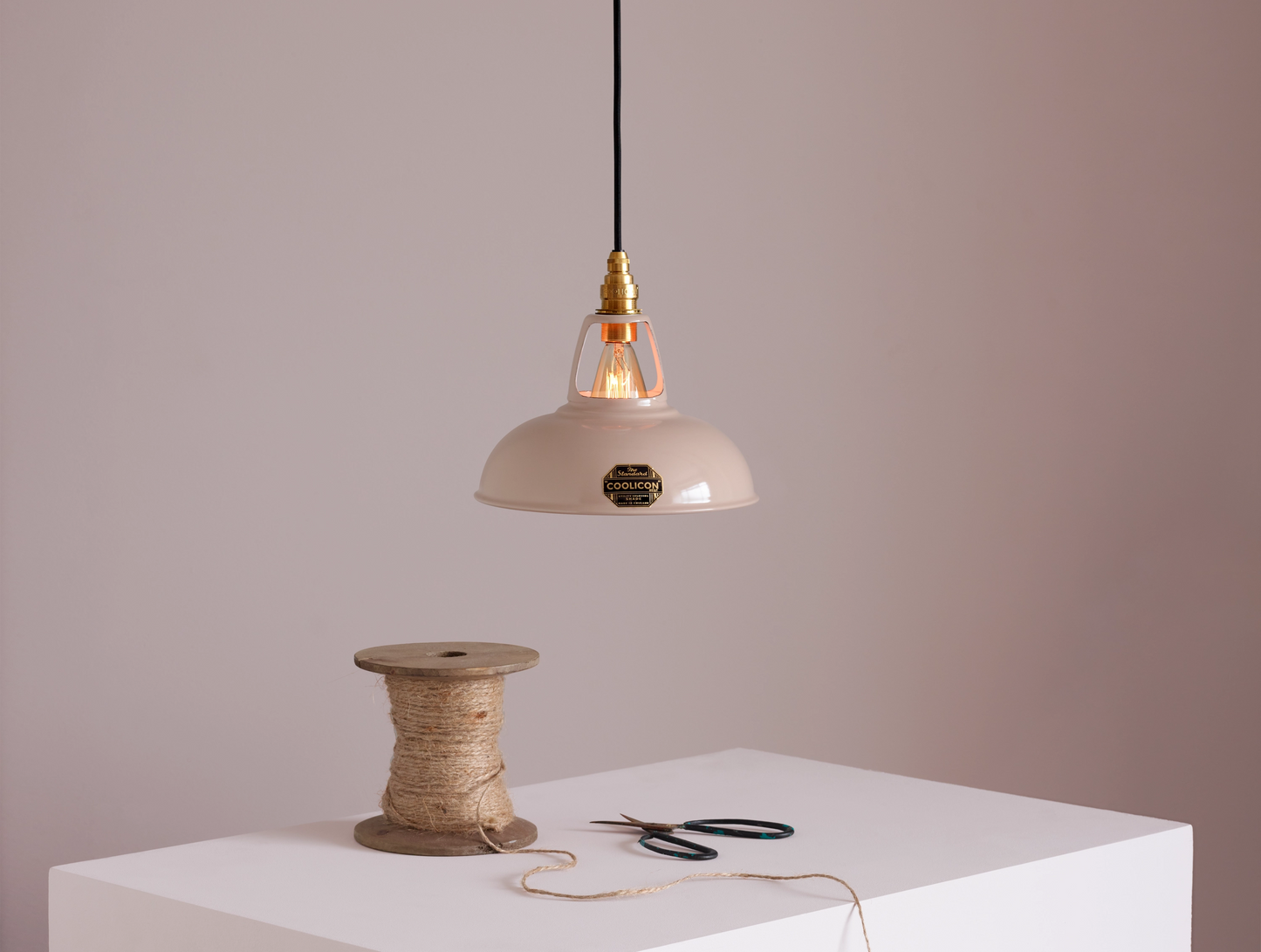 A Coolicon Latte Brown lampshade with a Signature Brass pendant set hanging above a plinth with a bobbin of twine and a pair of scissors