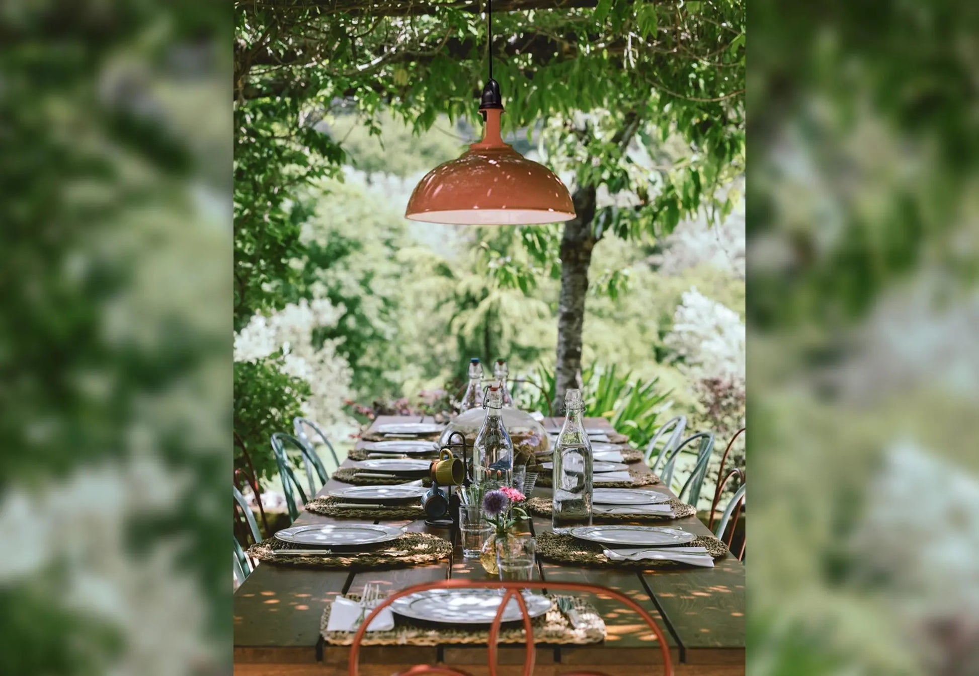 A Large Terracotta lampshade hanging from an outdoor suspension set, above a shaded table outside, surrounded by trees