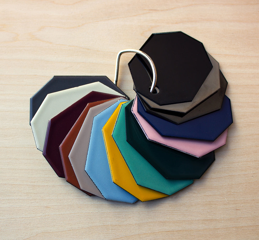 A Coolicon Lighting Swatch set displaying 16 Vitreous Enamel colours on a brass loop ring lying on a plywood surface.