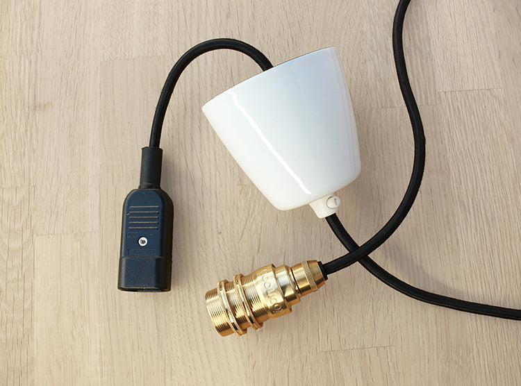 A Coolicon Lighting E14 Brass suspension set with black over braided cable and a white Vitreous Enamel Ceiling cup, with an IEC Connector on the other end. Product is laying on a plywood surface.