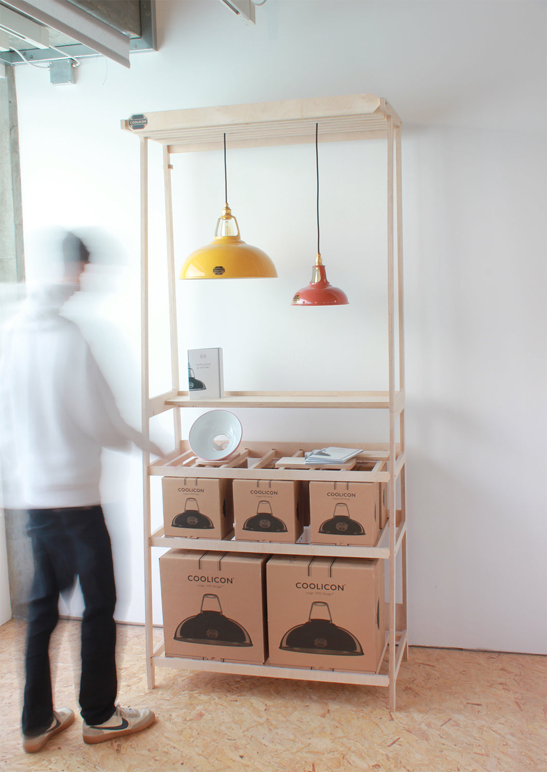 Plywood frame stand with two Coolicon Lampshades hanging and cardboard product boxes stacked on the lower shelves. A man is obscured in the foreground reaching for a product.