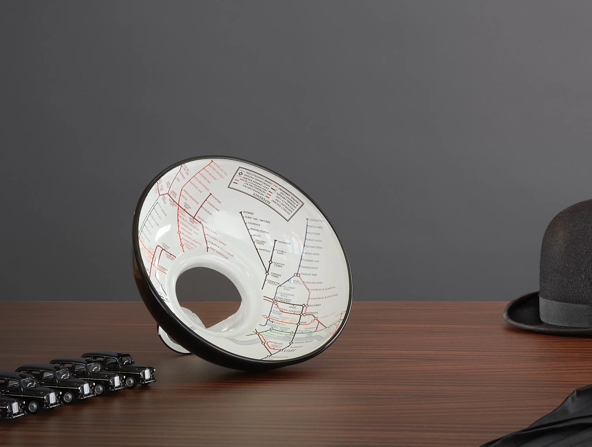 An Original Northern Line shade on a wooden table. On its left is a line of toy cars of black taxis. On the right is a black bowler hat and a black umbrella