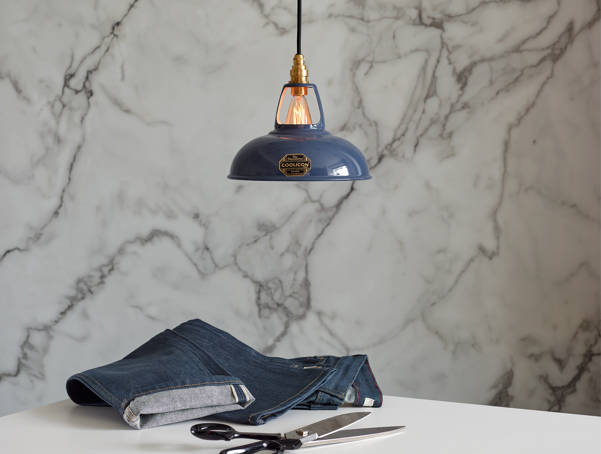 A Coolicon Selvedge blue lampshade with a Signature Brass pendant set hanging above a table with a pair of jeans and fabric scissors. The background is a marble wallpaper.
