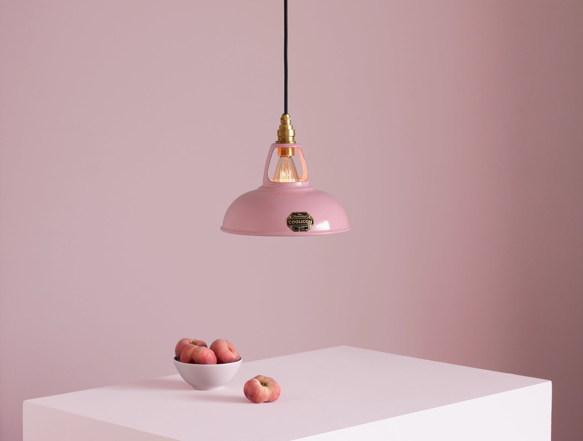 A Coolicon Powder Pink lampshade with a Porcelain pendant set hanging above a plinth with a bowl of flat peaches.