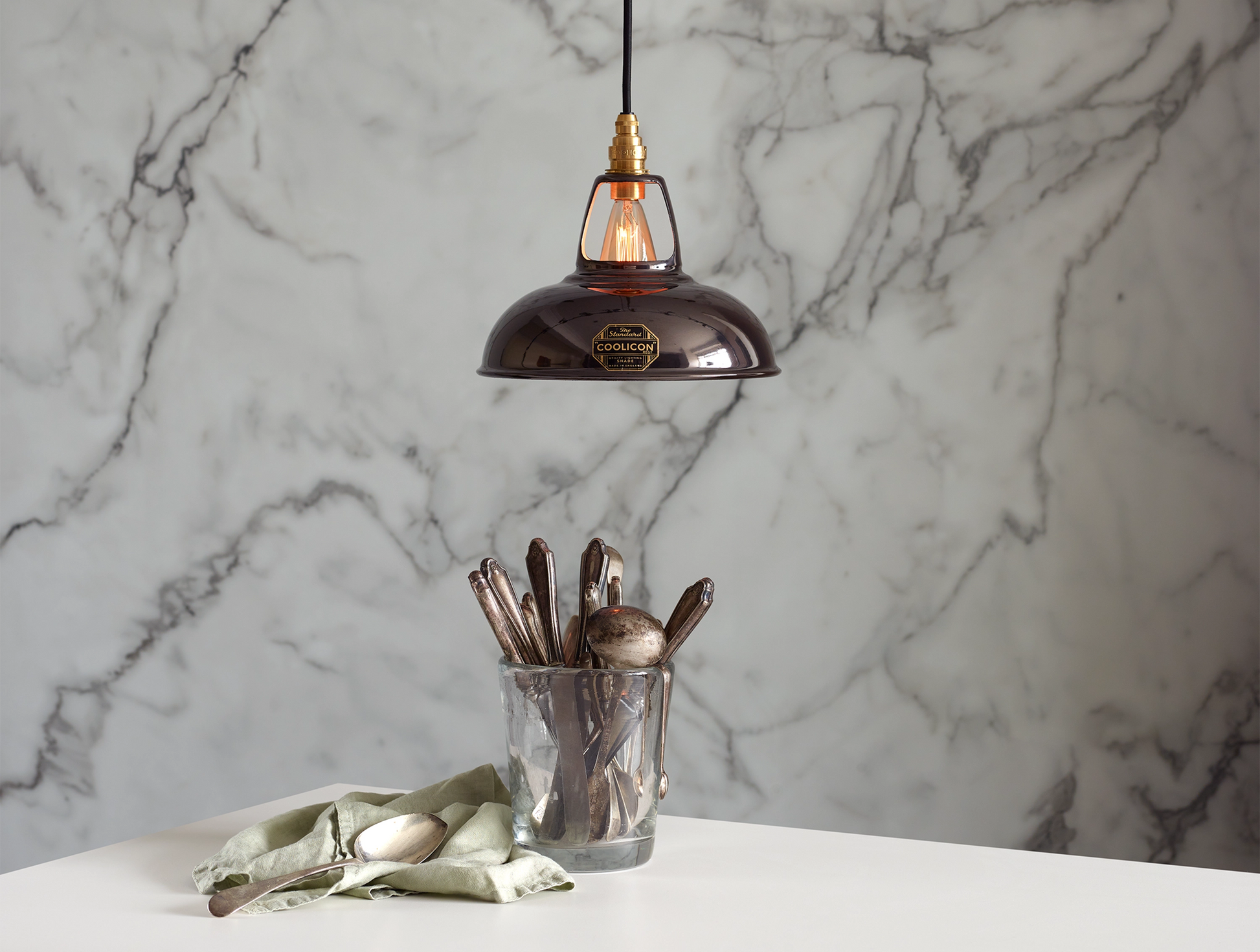 A Coolicon Pewter grey lampshade with a Signature Brass pendant set hanging above a table with a glass container with a set of old cutlery. The background is a marble wallpaper.