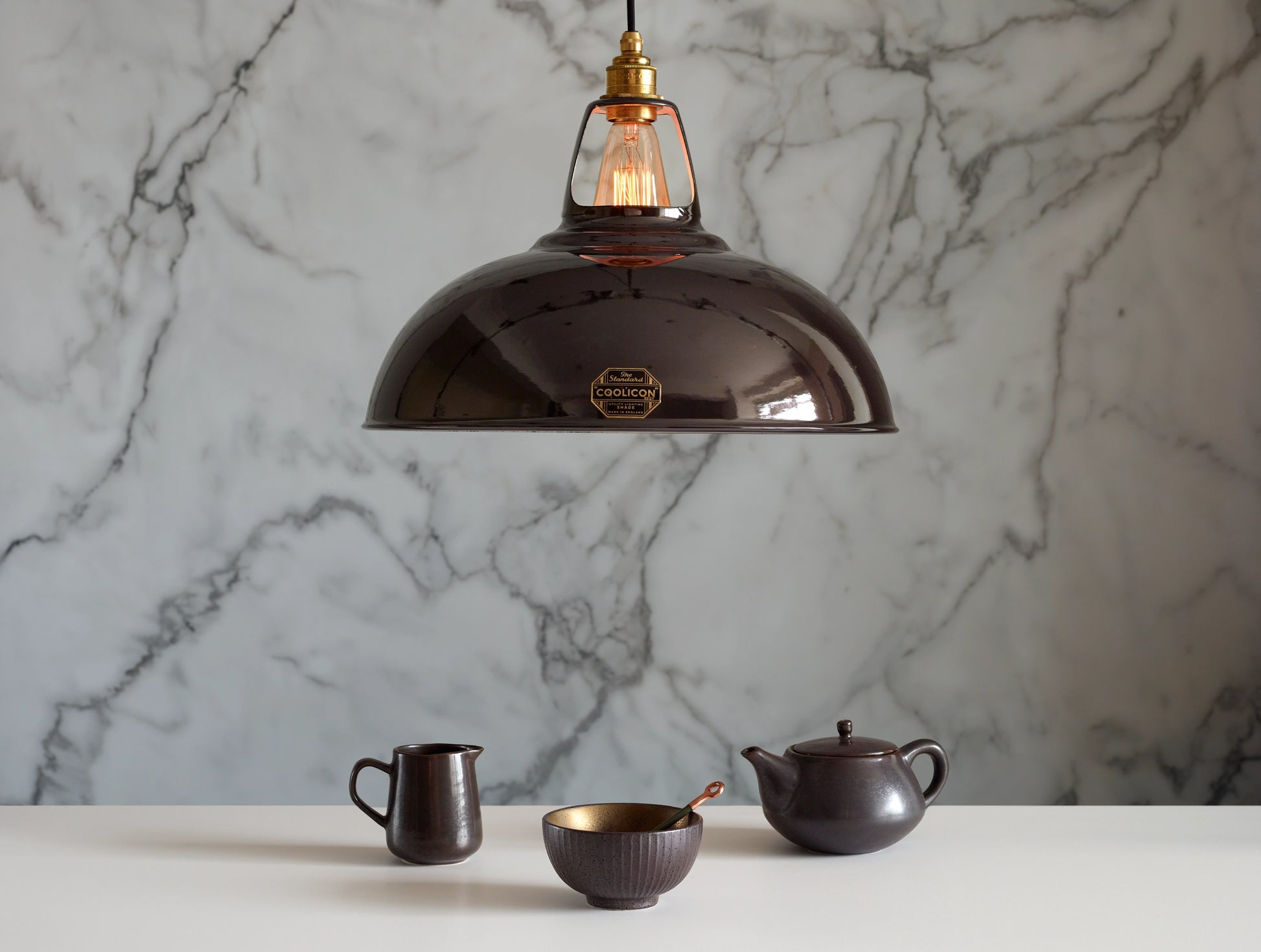 A Large Coolicon Pewter grey lampshade with a Signature Brass pendant set hanging above a table with a matching set of dark tea accessories. There is a milk jug, a matcha bowl with a copper spoon and a teapot