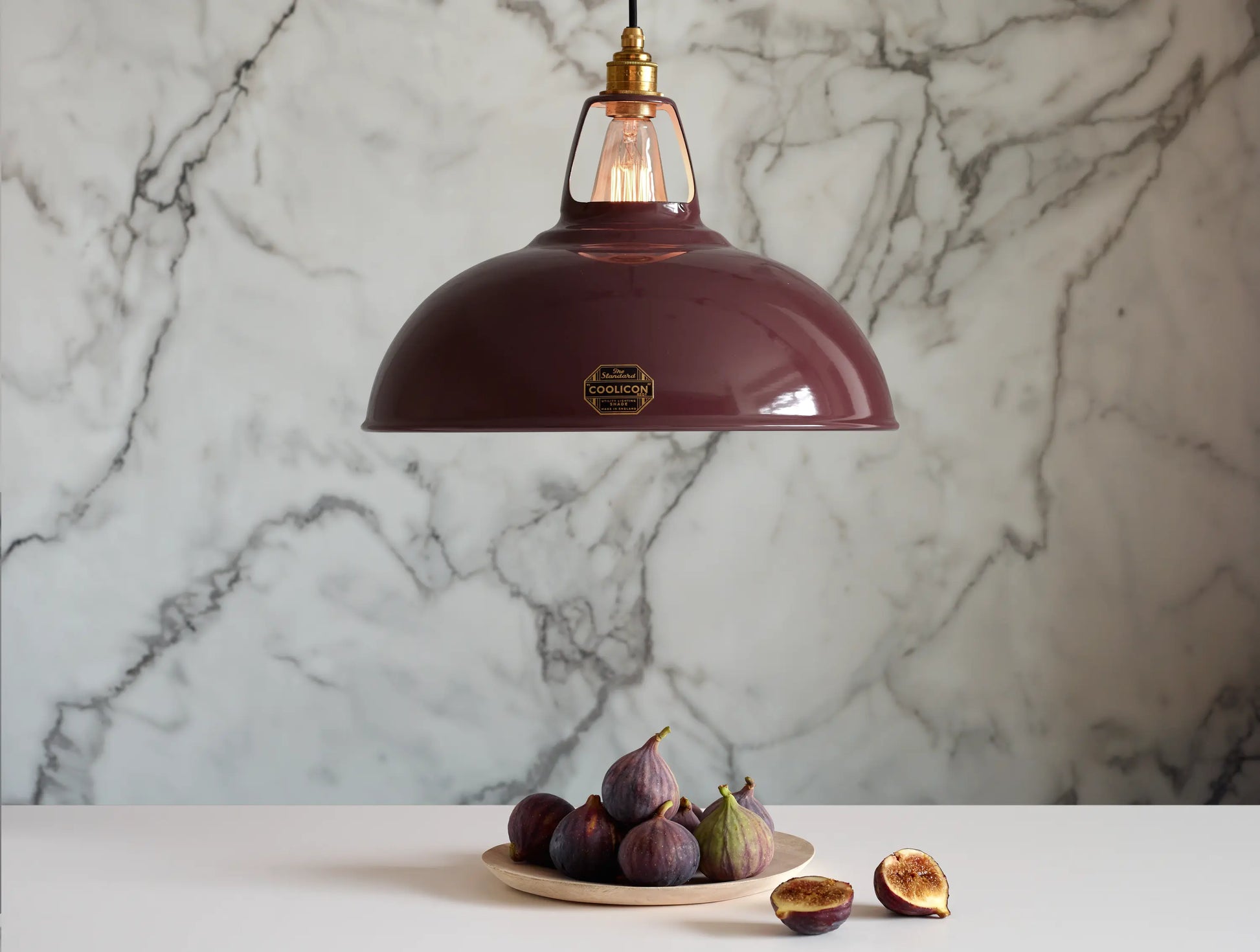 A Large Coolicon Metropolitan burgundy lampshade with a Signature Brass pendant set hanging above a table with tray of figs. One fig has been cut in half. The background is a marble wallpaper.