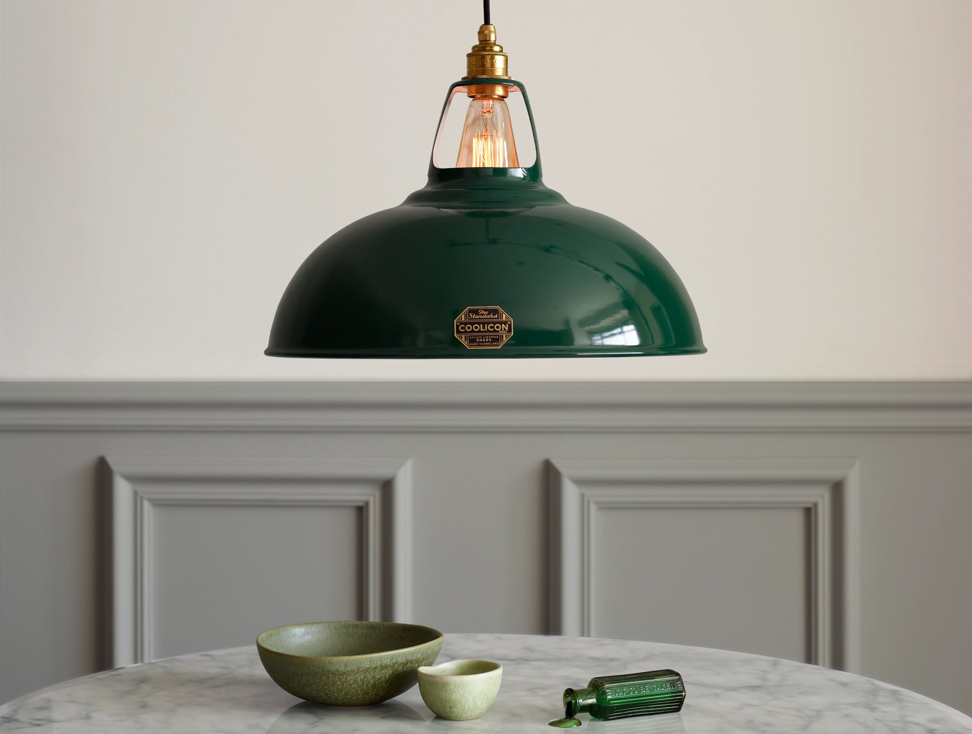 A Large Coolicon Original Green lampshade hanging over a marble table. Two green bowls and a small green glass bottle spilling a green liquid are placed on the table