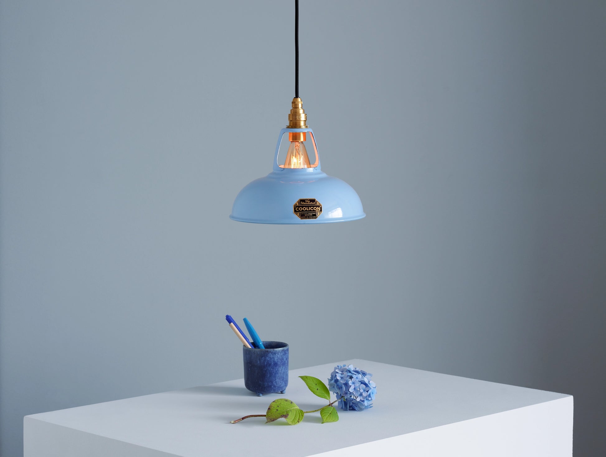 A Coolicon Sky Blue lampshade with a Porcelain pendant set hanging above a plinth with a blue hydrangea flower and a dark blue pencil cup containing two blue pens