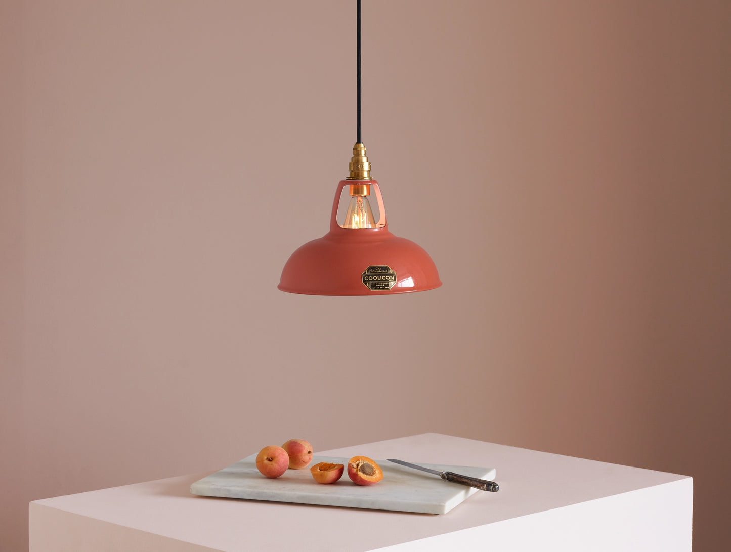 A Coolicon Terracotta lampshade hanging over a plinth. Below the shade is a marble cutting board with a knife and three apricot. One of them is cut in half