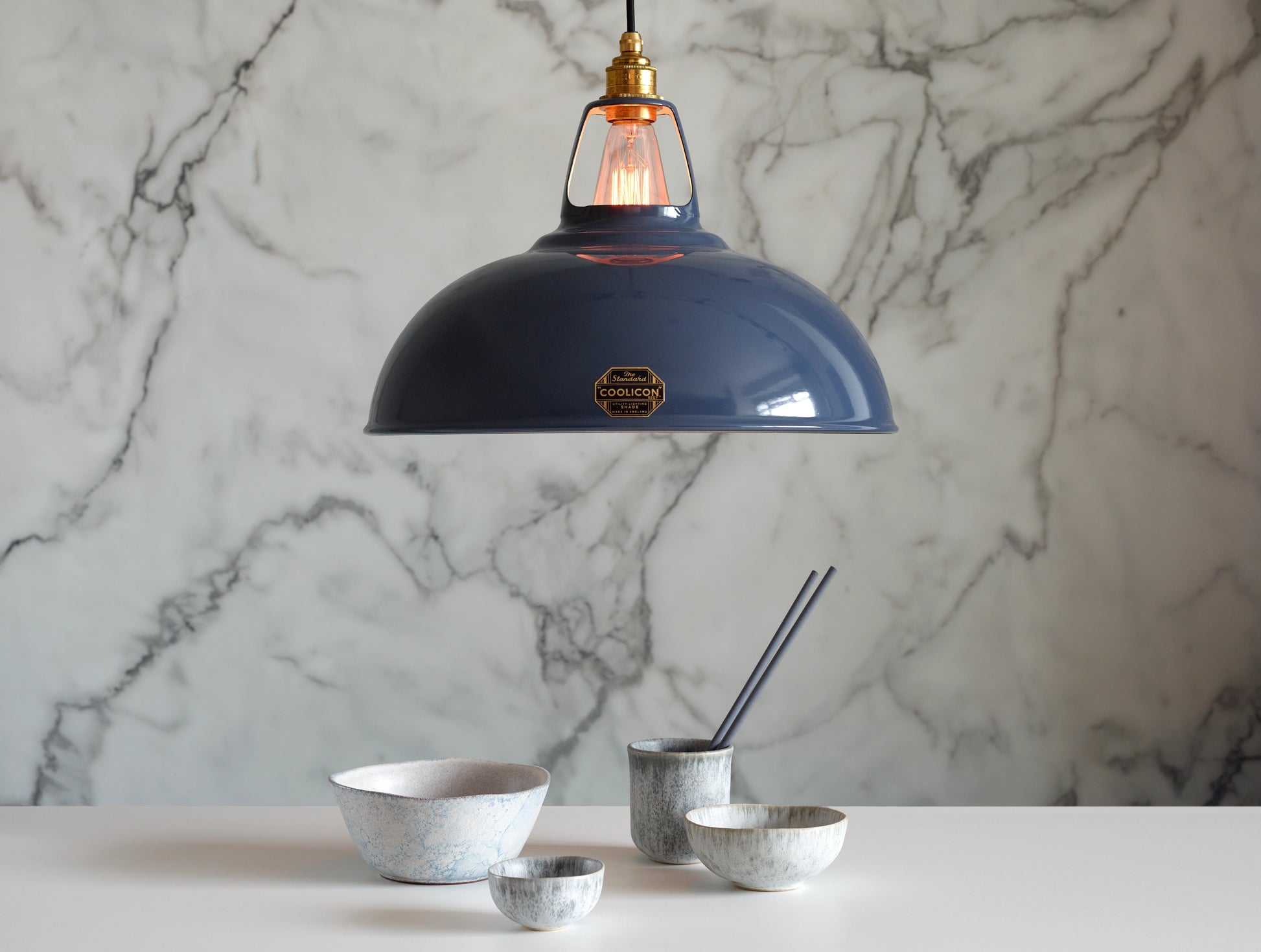 A Large Coolicon Selvedge blue lampshade with a Signature Brass pendant set hanging above a table with a set of white and blue bowls of different sizes and a pair of blue chopsticks. The background is a marble wallpaper.