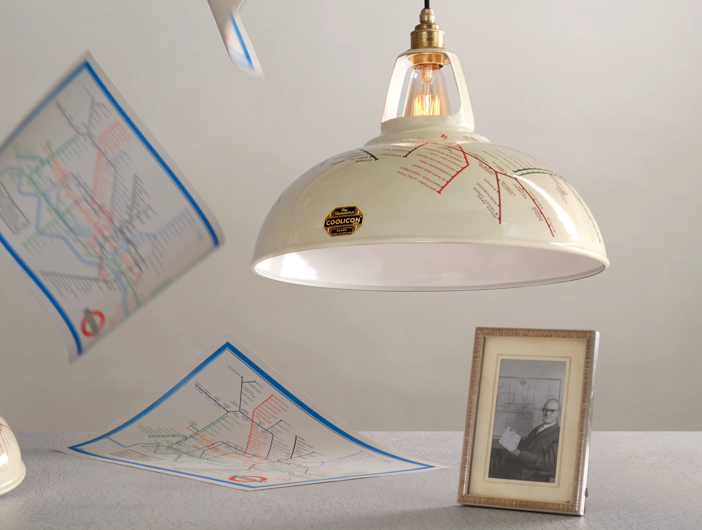 A Large Underground Map shade above a table. There is a framed black and white portrait of Harry Beck on the table. In the background, three 1933 maps are falling from the ceiling